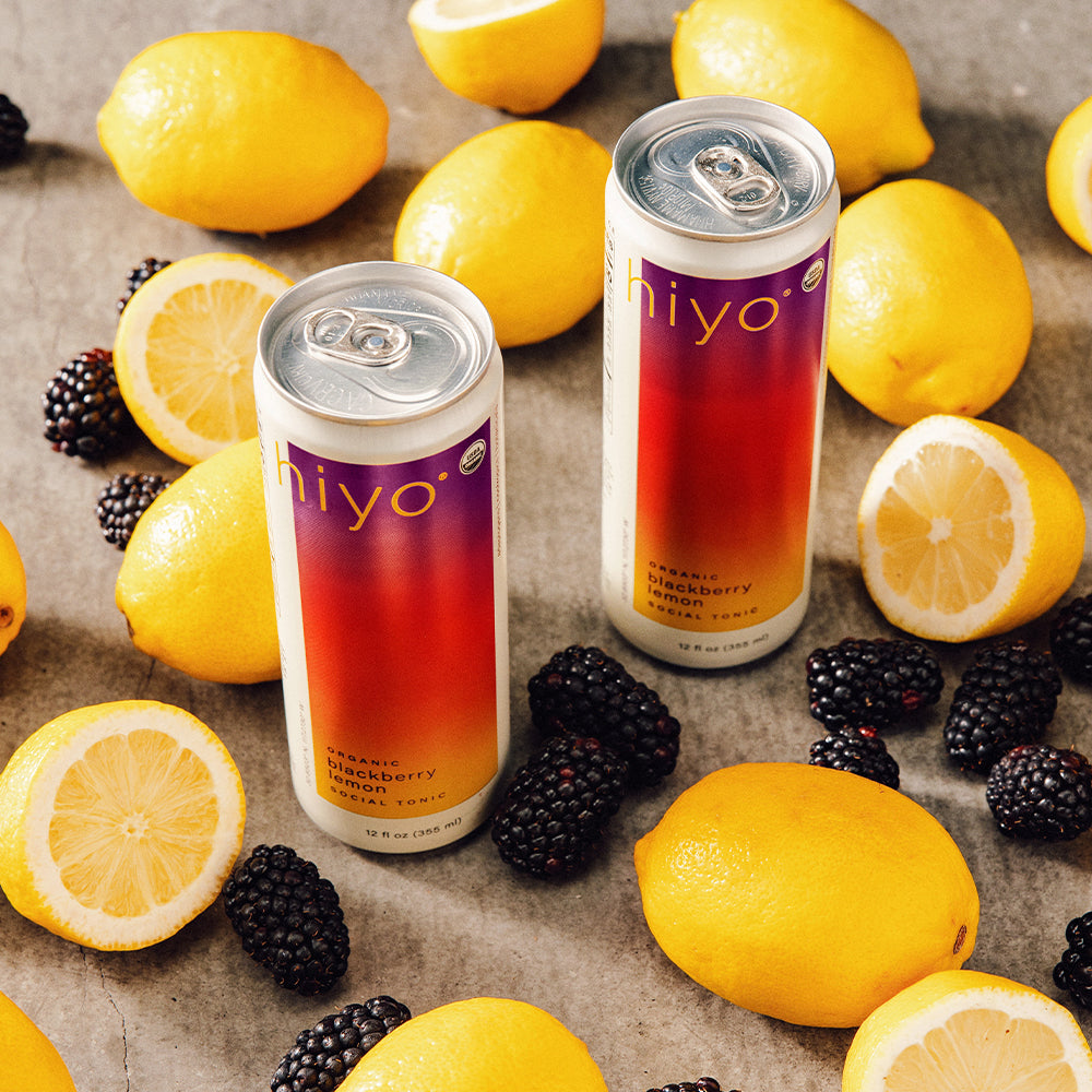 Two cans of non-alcoholic functional seltzer hiyo blackberry lemonade surrounded by lemons and blackberries on a textured surface.