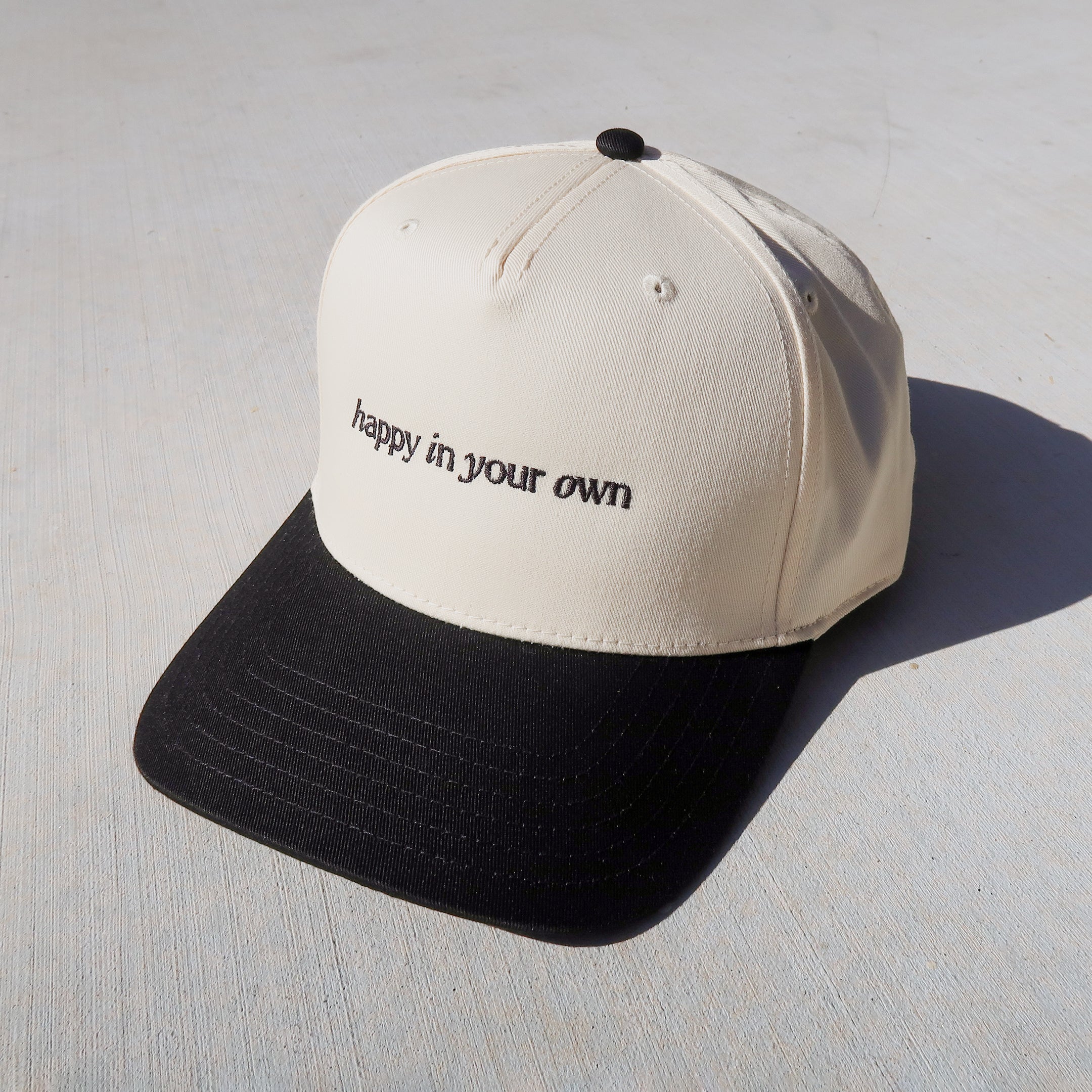 a two-tone trucker hat featuring a black brim and ivory body. it includes the text 'happy in your own' embroidered across the front center in black thread.