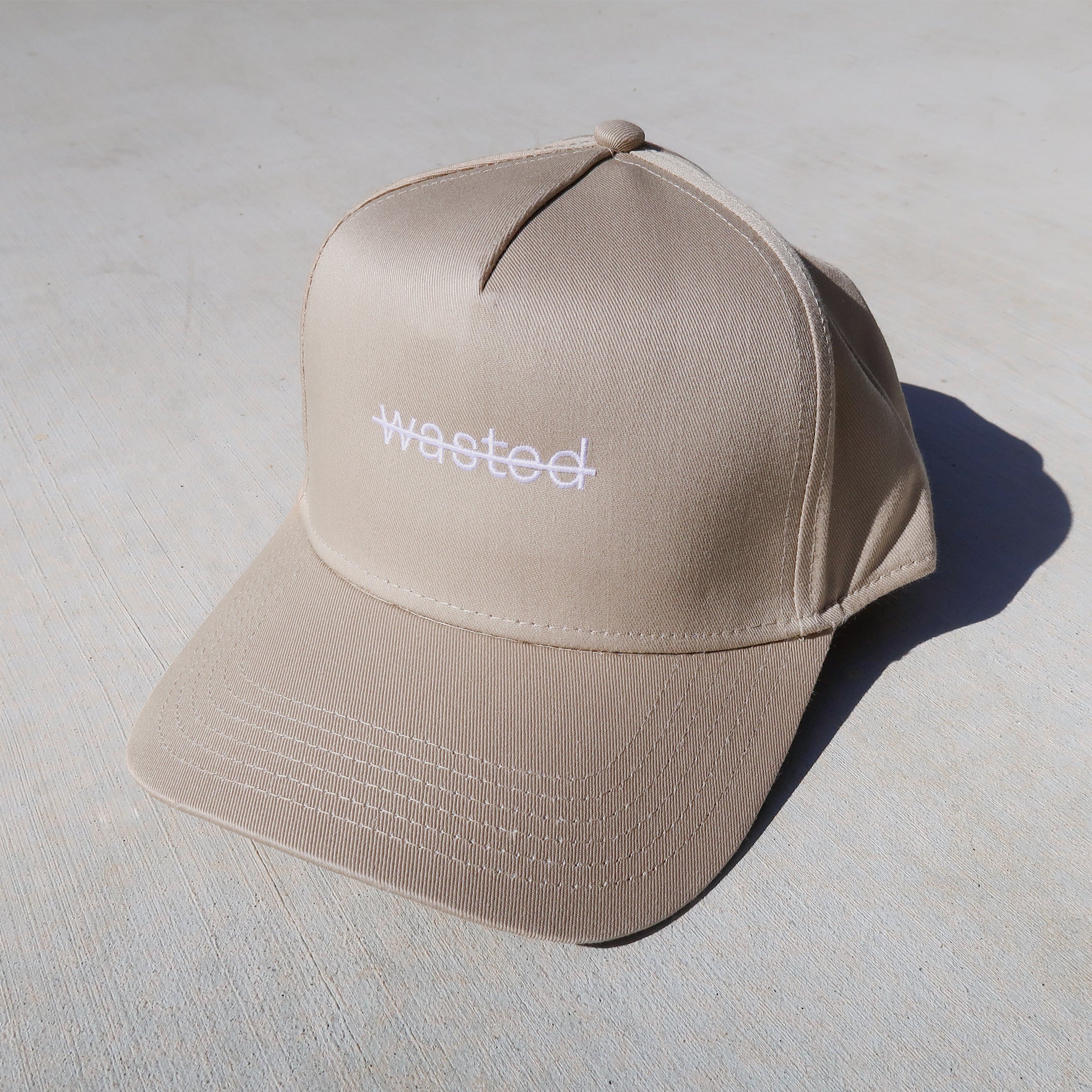 a beige trucker hat featuring that includes the text 'wasted' struck through and embroidered across the front center in white.