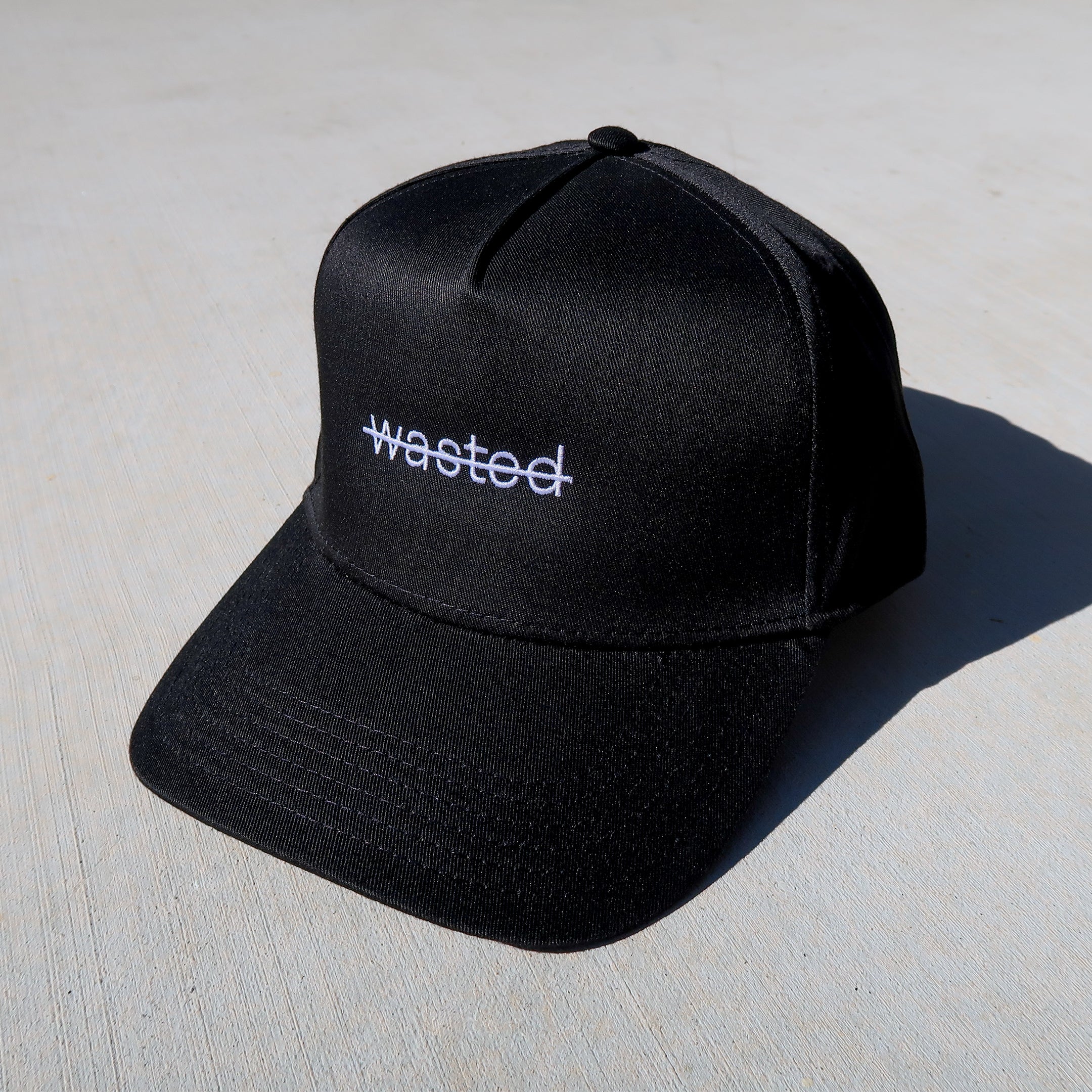 a black trucker hat featuring that includes the text 'wasted' struck through and embroidered across the front center in white.