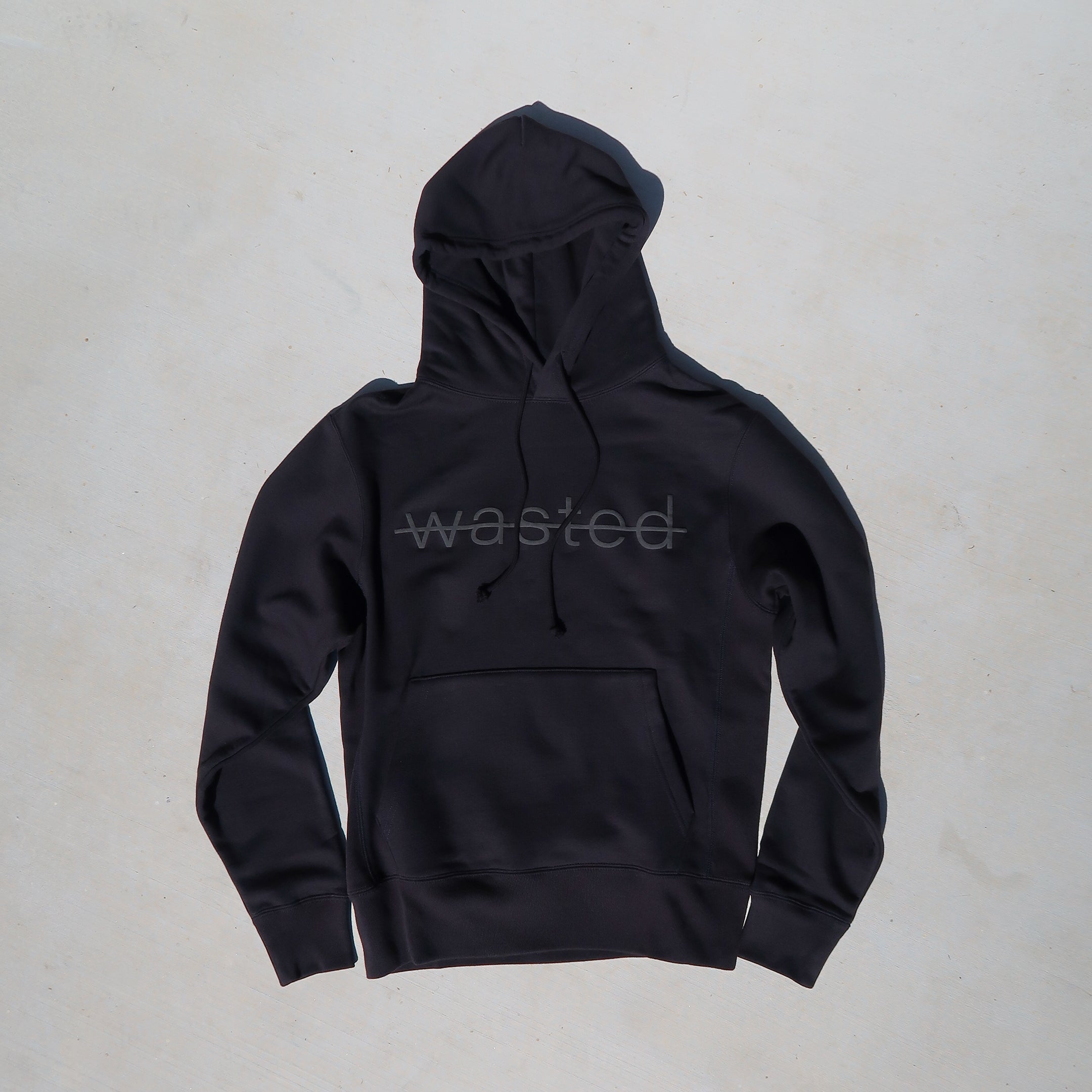 black drawstring hoodie with the text 'wasted' struck through and printed across the front chest in black puff ink.