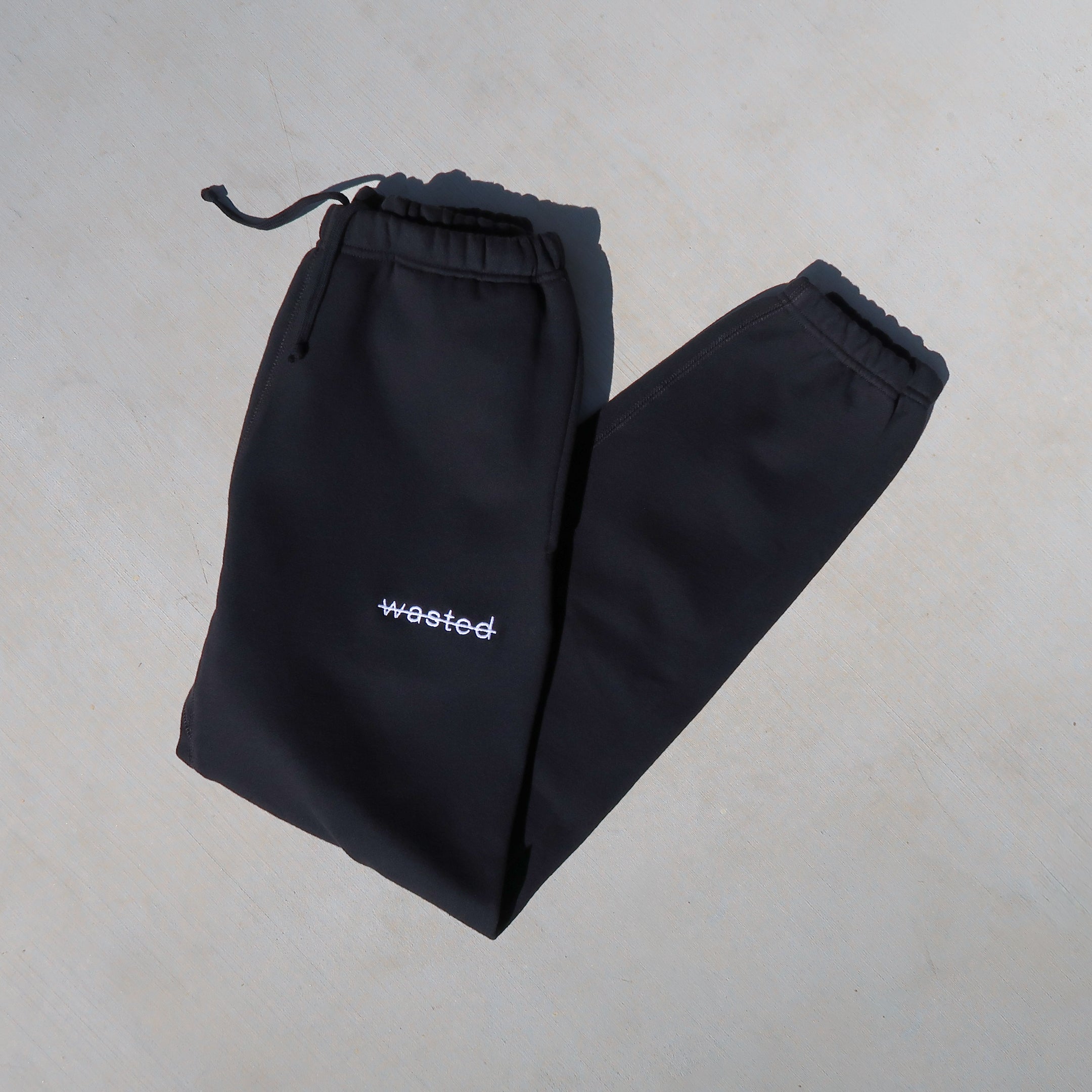 black drawstring sweatpants with the text 'wasted' struck through and embroidered onto the front left hip in white.