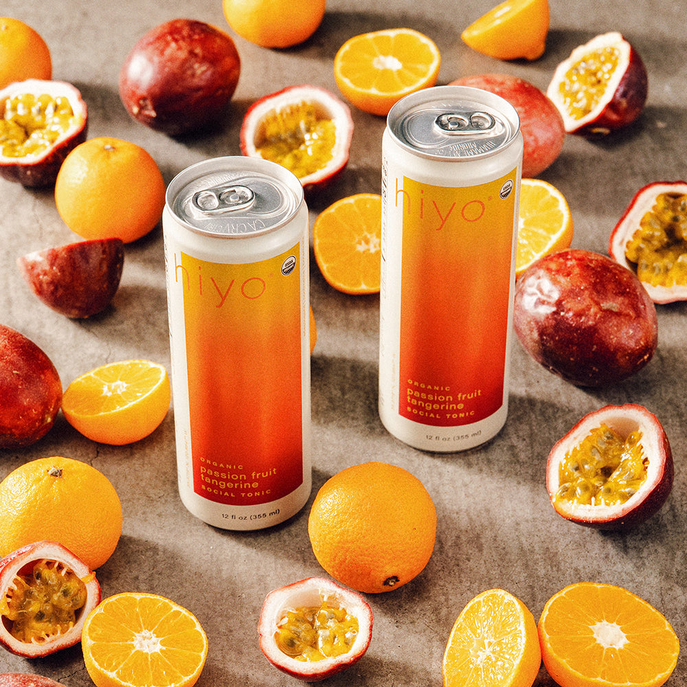 Two cans of non-alcoholic hiyo passion fruit tangerine on a surface with oranges and passion fruits.