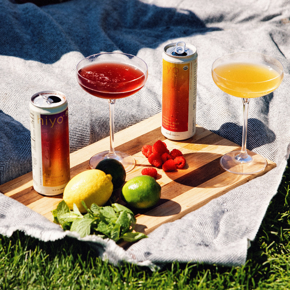 Two mocktails in glasses with cans of non-alcoholic functional seltzer hiyo on a wooden board outdoors.