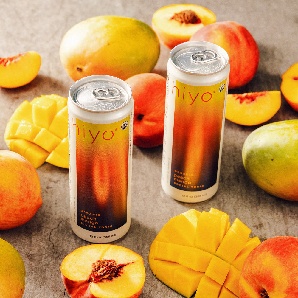 Two cans of non-alcoholic functional seltzer hiyo organic peach mango fruit tonics surrounded by sliced peaches and mangoes on a stone surface.