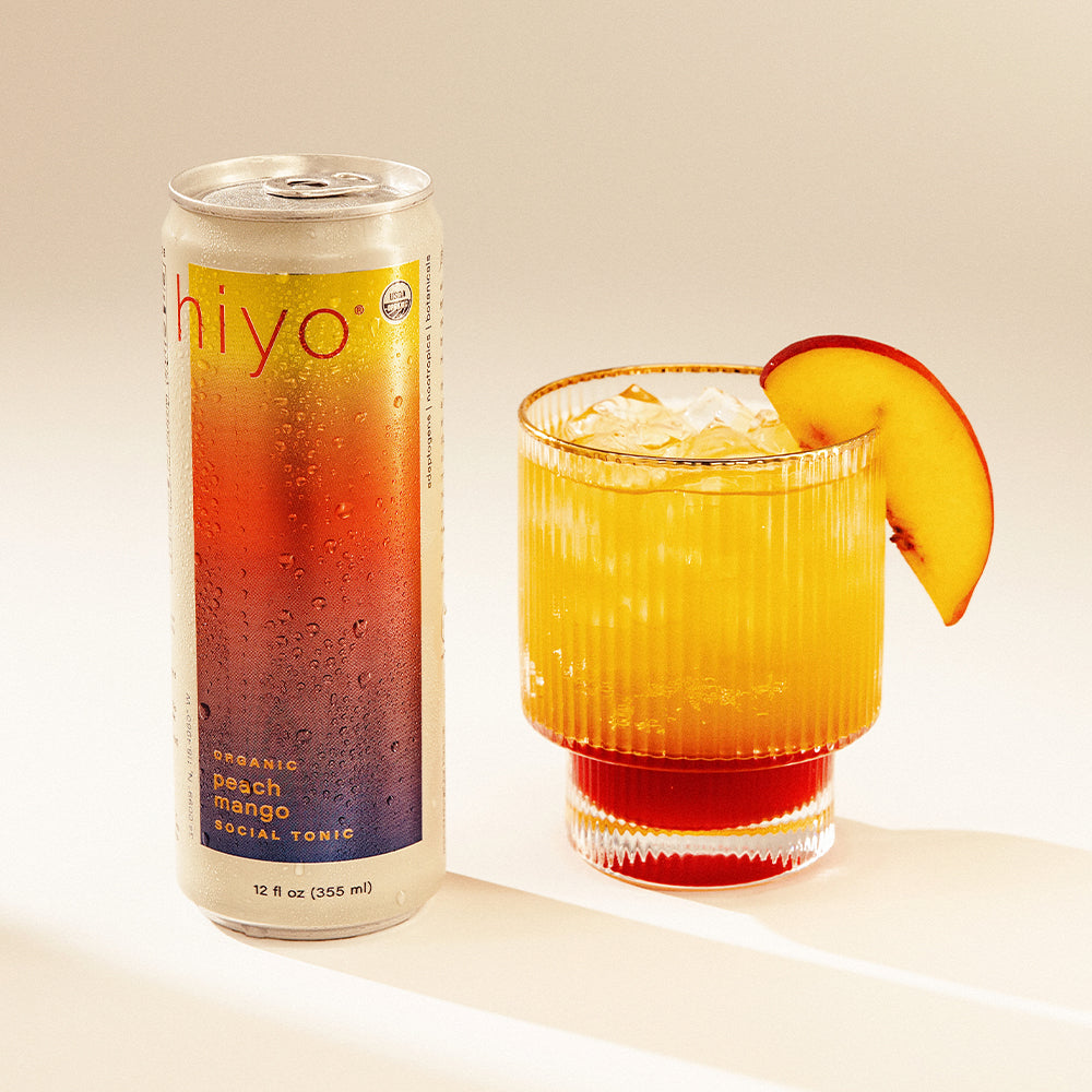 Can of non-alcoholic functional seltzer hiyo peach mango social tonic next to mocktail in a glass with a peach slice on a beige background