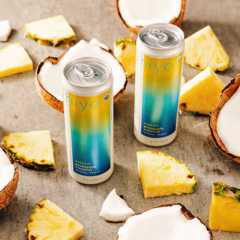 Two cans of non-alcoholic functional seltzer hiyo drink surrounded by pineapple and coconut pieces on a textured surface.
