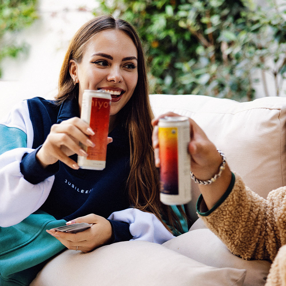 Two people sitting on a couch, each holding a can of non-alcoholic functional seltzer hiyo with a blurred background