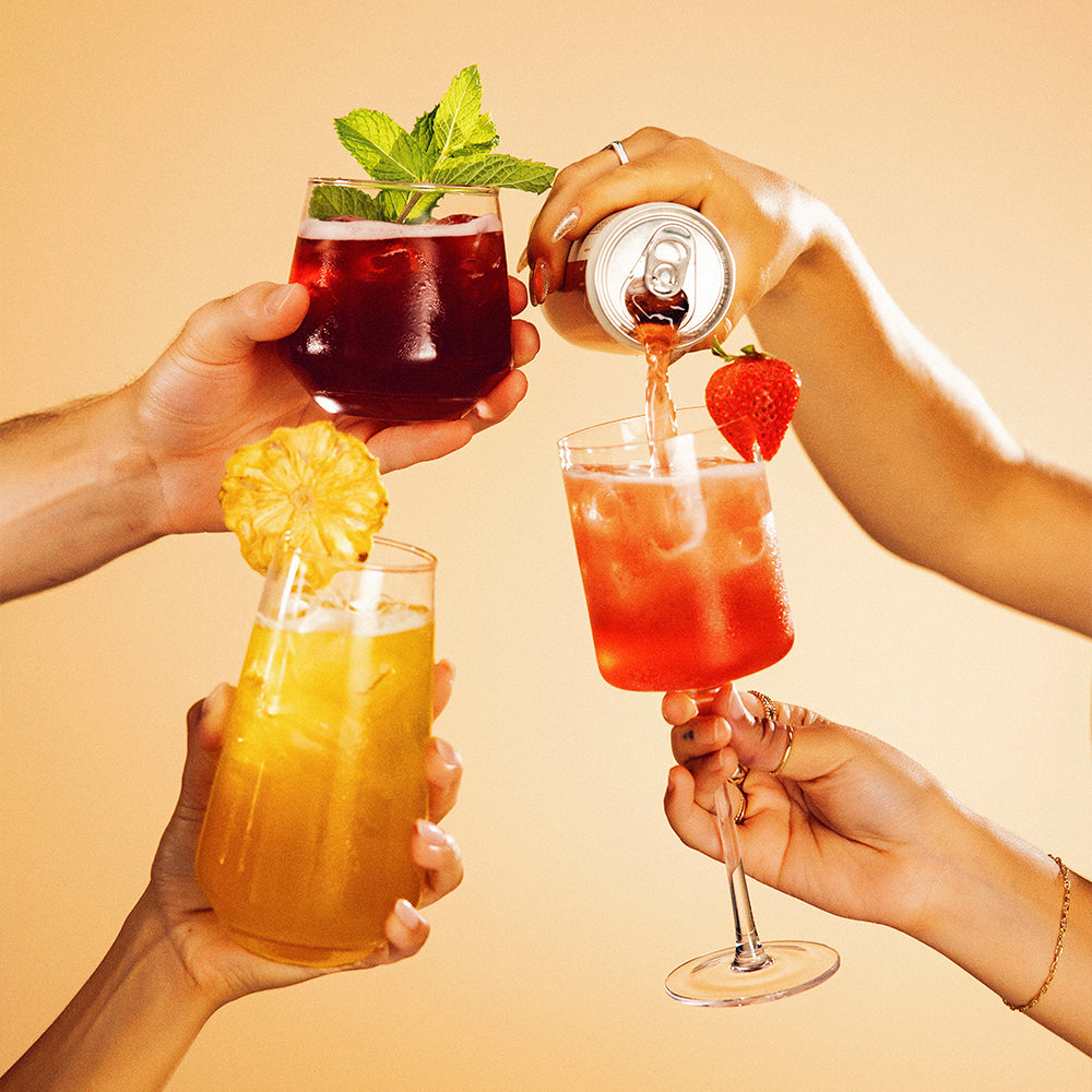 Four hands holding non-alcoholic functional seltzer hiyo mocktails with a can being poured into one of them against a beige background
