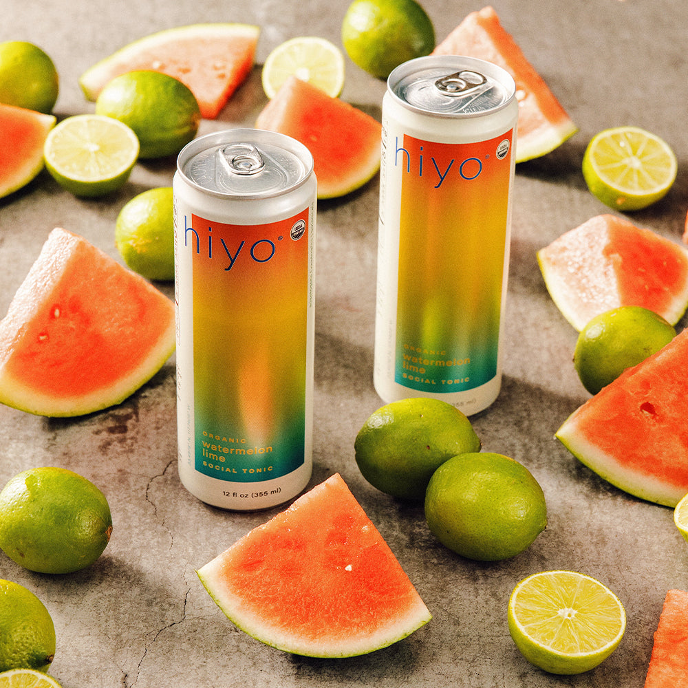 Two cans of non-alcoholic functional seltzer hiyo watermelon lime surrounded by watermelon slices and limes on a textured surface.
