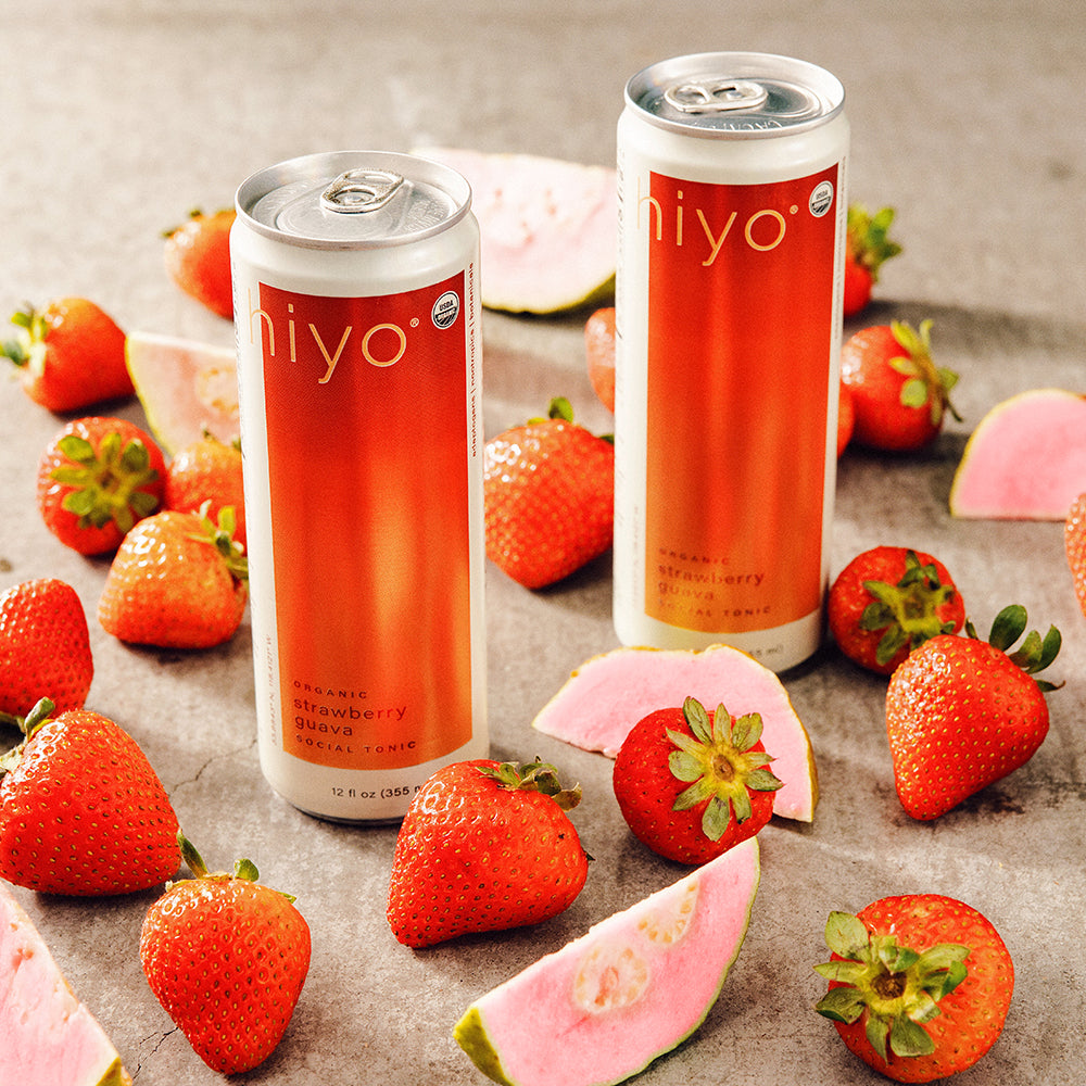 Two cans of non-alcoholic hiyo strawberry drink surrounded by strawberries and guava slices on a textured surface.