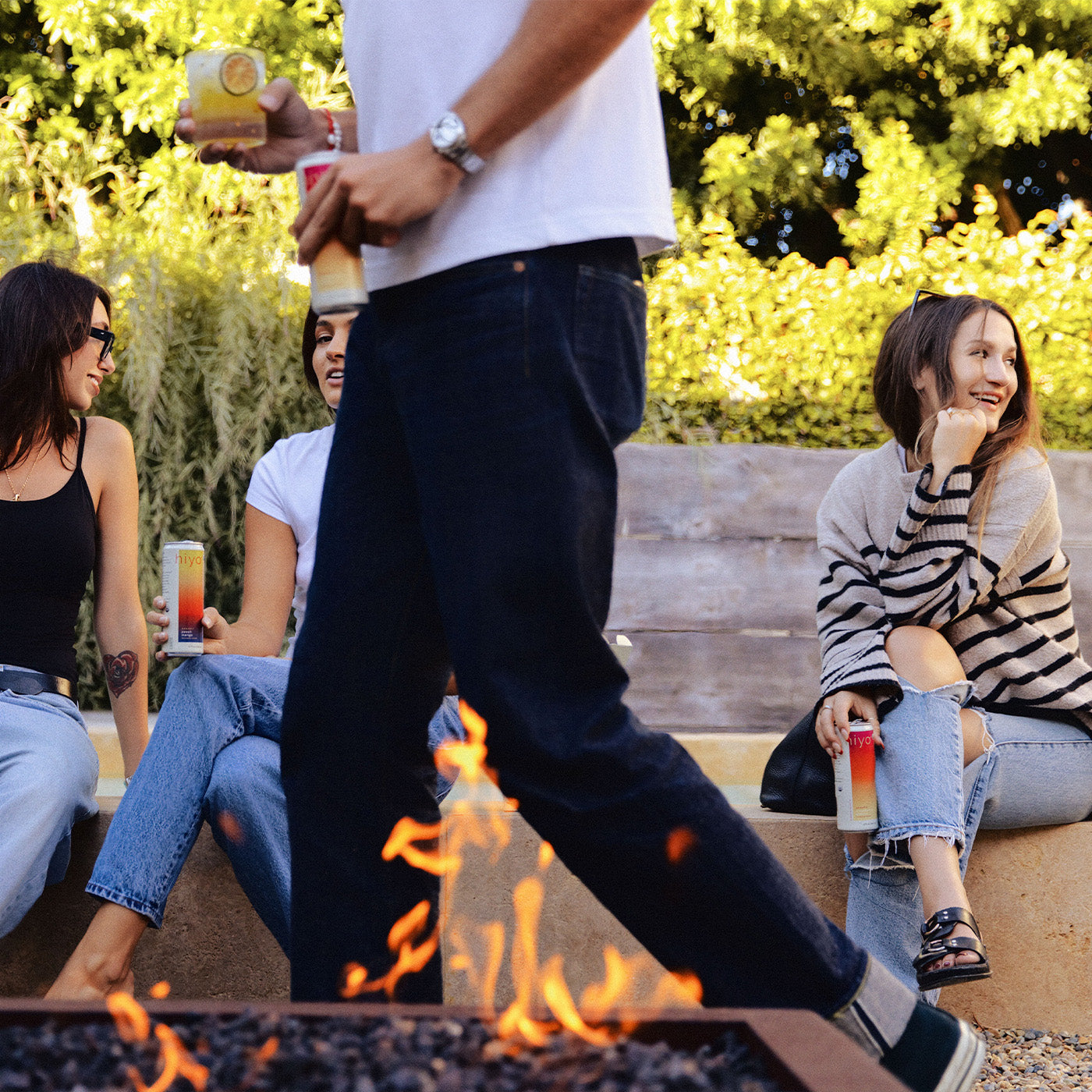 a group of friends sitting next to a bonfire with cans of hiyo and mixed drinks in hand.