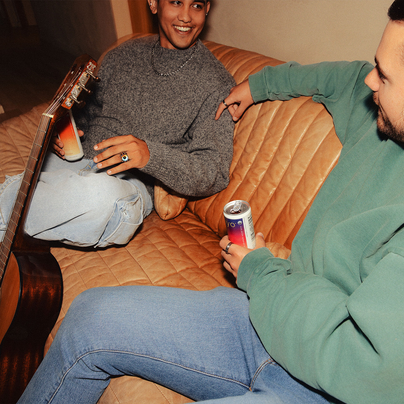 two men sitting together with hiyo in hand on a leather couch with a guitar beside it.