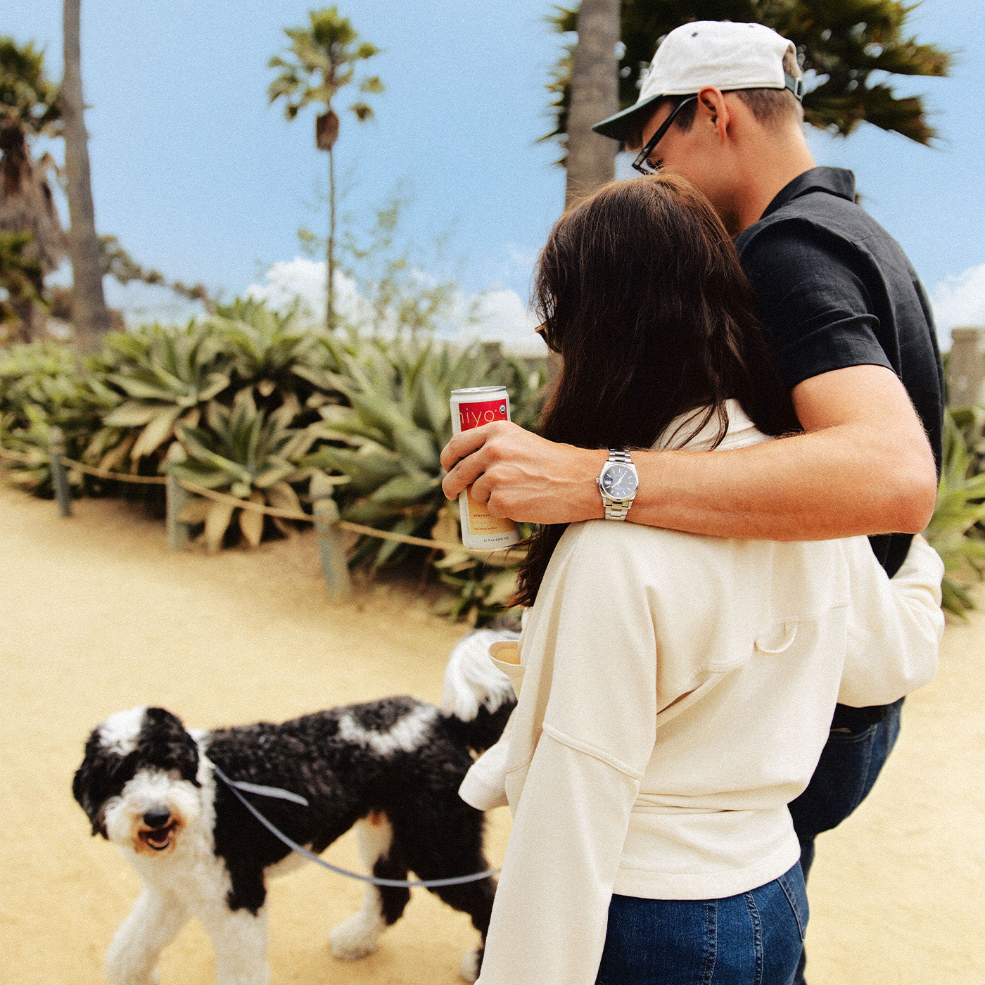 a man and woman walk their dog along the a tropical pathway while embraced. the man is holding a can of strawberry guava hiyo over the woman's shoulder.