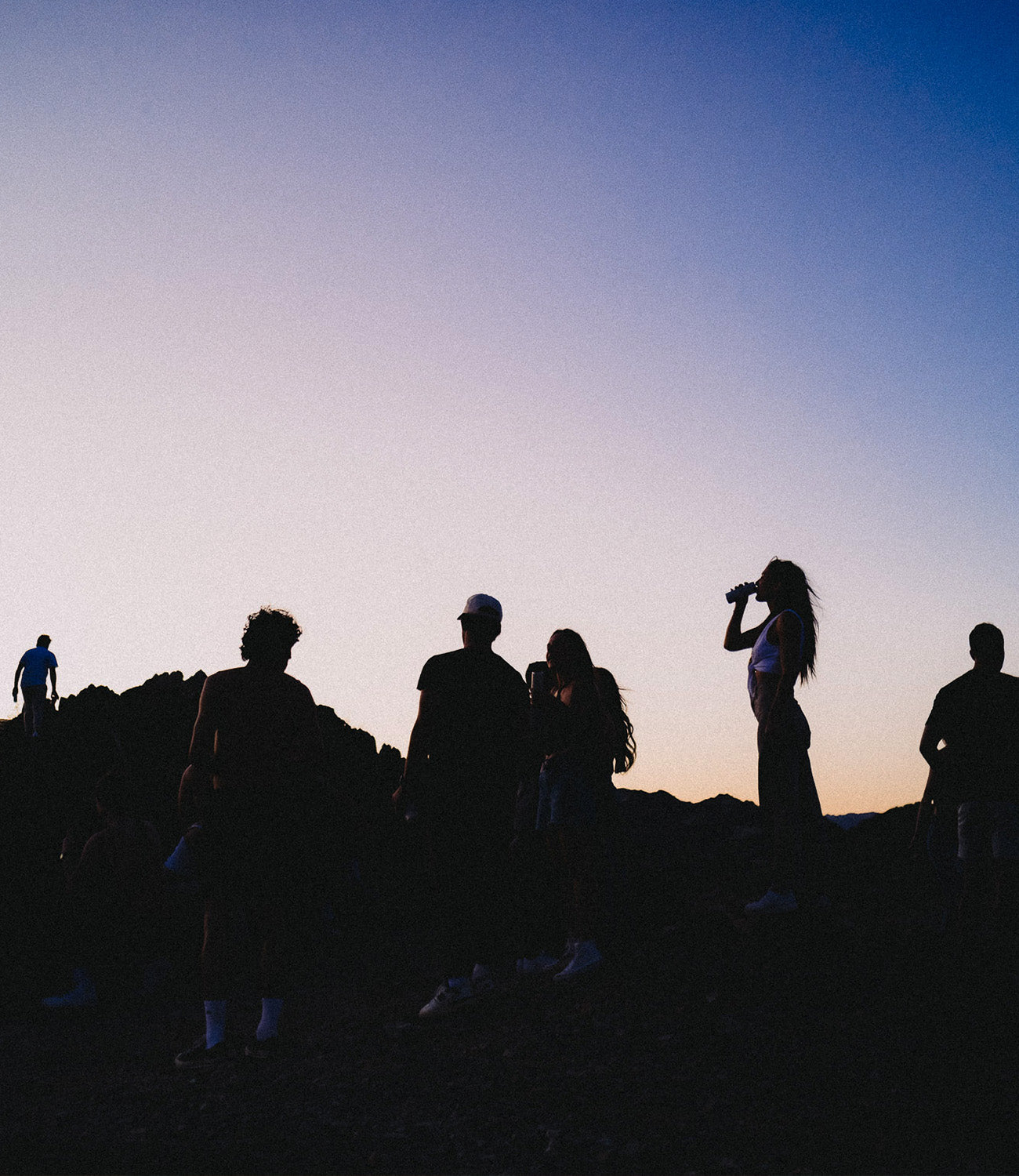 a group of people standing in the desert while the sun sets to the horizon. a woman sips a hiyo while others explore.