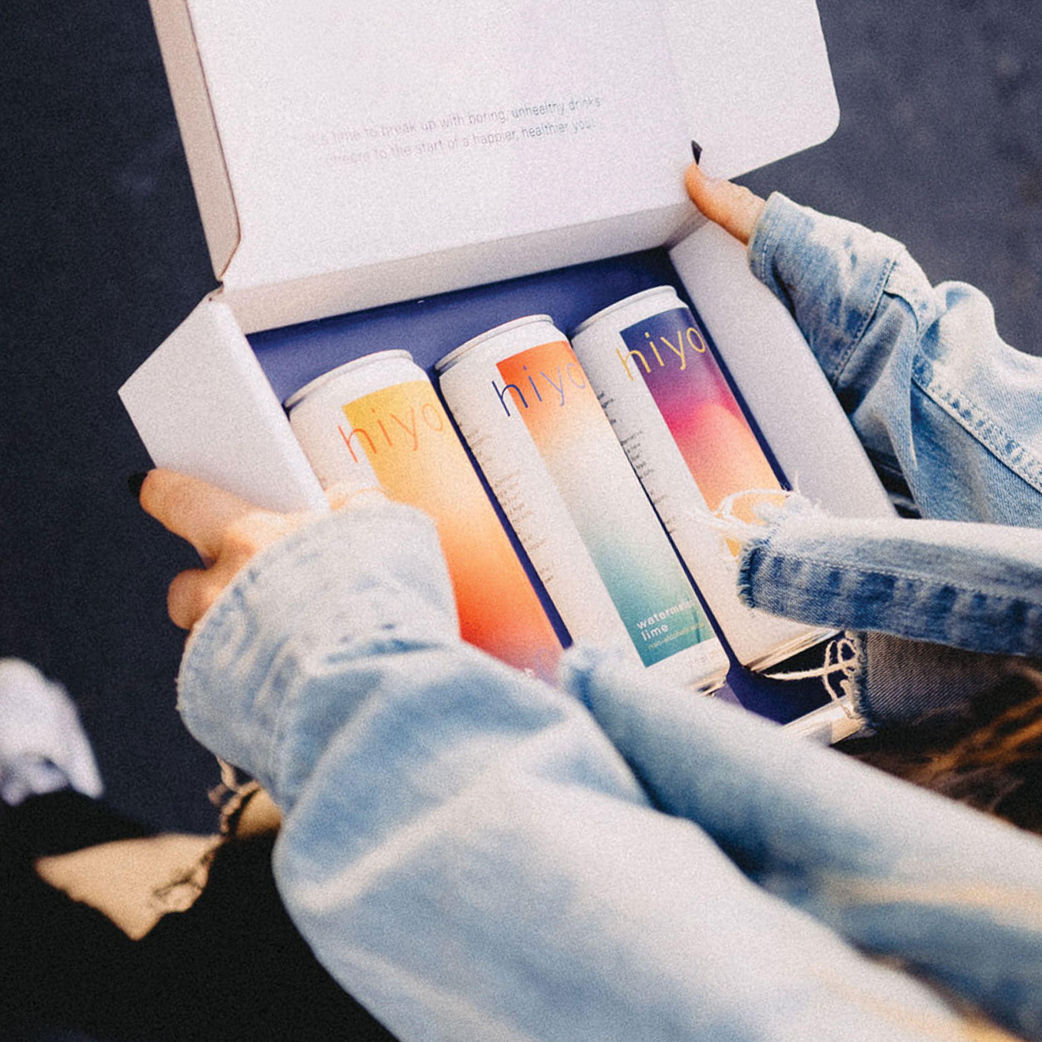 a woman in a jean jacket holding a sampler box which contains three cans of hiyo.