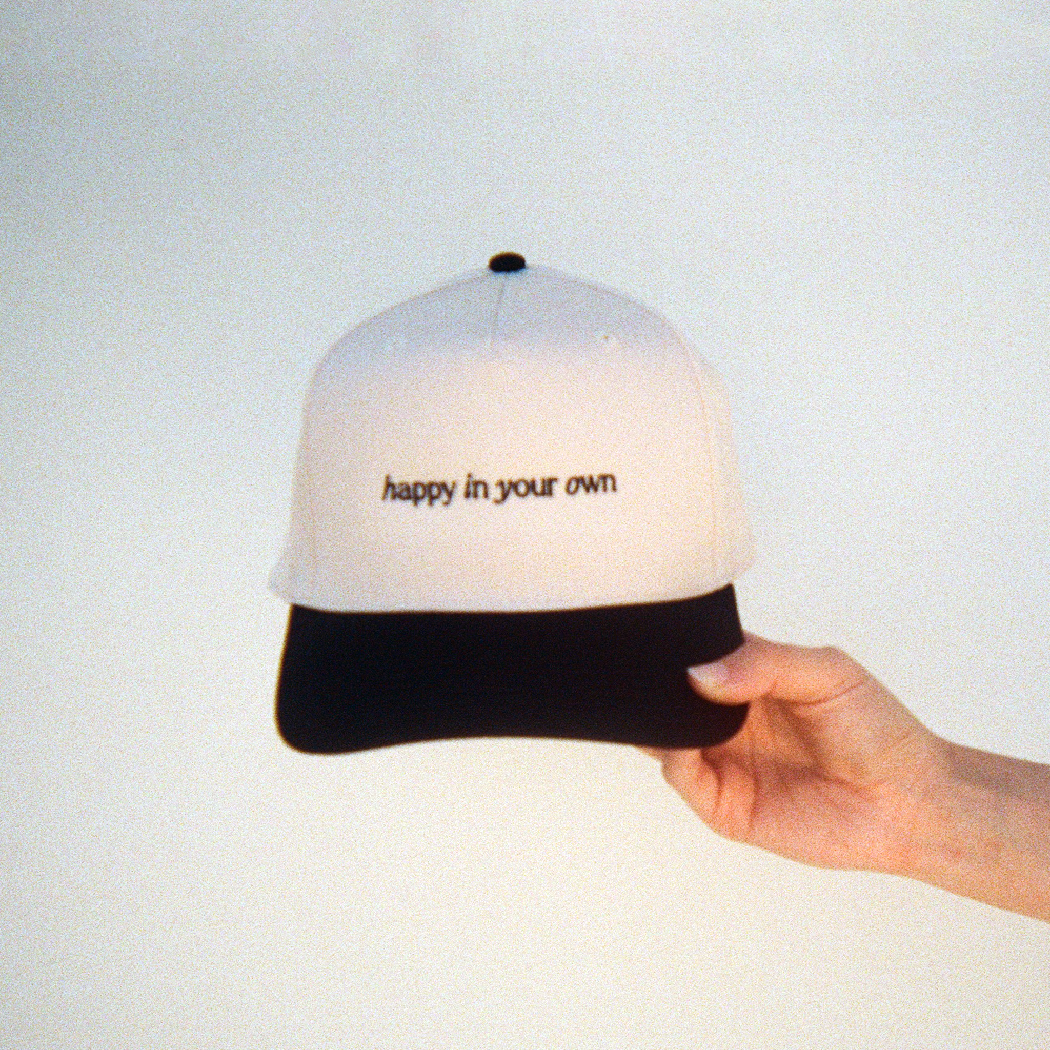 a two-tone trucker hat featuring a black brim and ivory body. it includes the text 'happy in your own' struck through and embroidered across the front center in black thread.