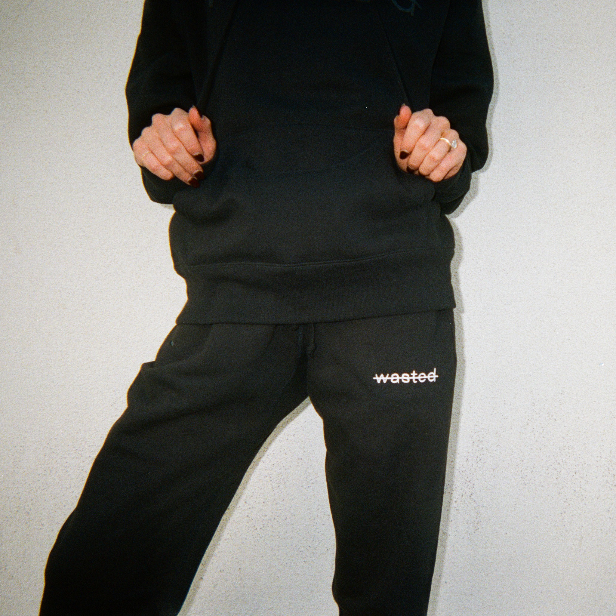 a woman wearing black drawstring sweatpants with the text 'wasted' struck through and embroidered onto the front left hip in white thread.