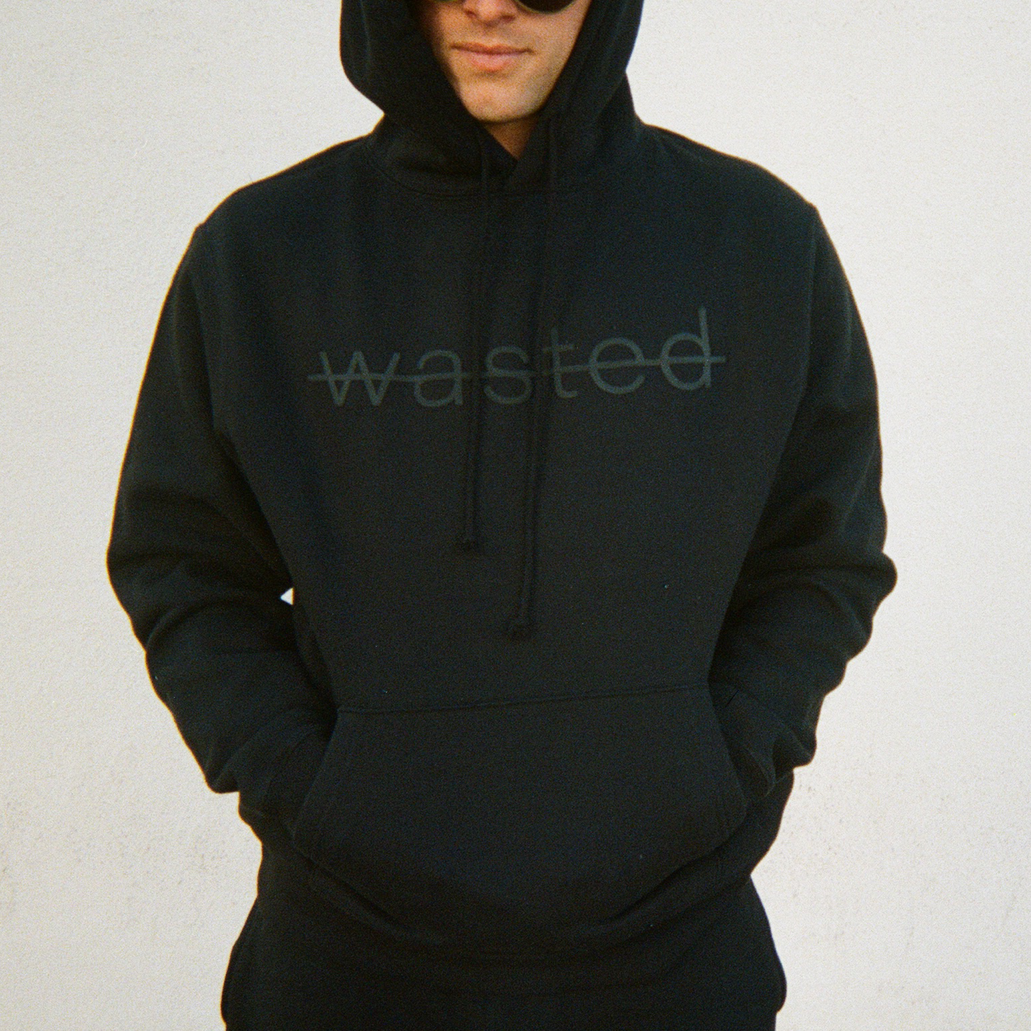 a man wearing a black drawstring hoodie with the text 'wasted' struck through and printed across the front chest in black puff ink.